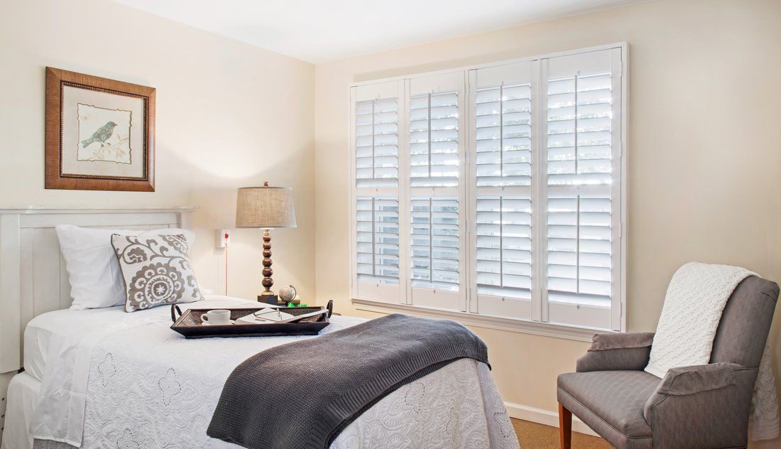 A cozy and well-lit bedroom featuring a bed with a tray, a bedside lamp, and large windows with shutters.