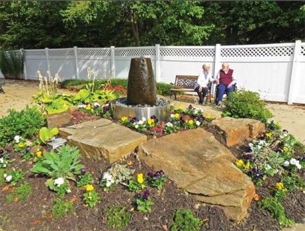 A beautifully landscaped garden with a central fountain, surrounded by flowers and large rocks. Two people are sitting on benches in the background, enjoying the serene setting enclosed by a white fence.