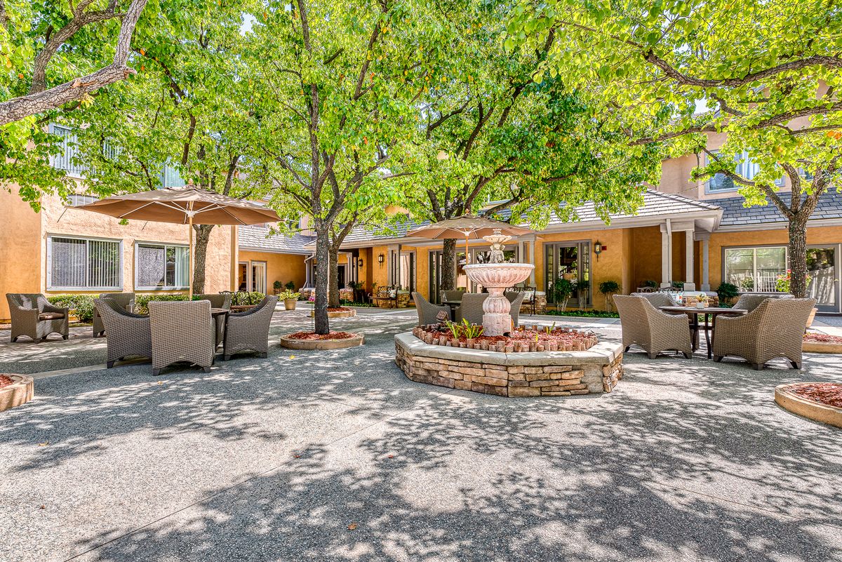 A tranquil courtyard featuring a central fountain surrounded by patio furniture under the shade of trees.
