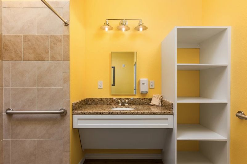 A bathroom with a yellow wall, featuring a vanity sink, mirror, shelves, and overhead lighting.