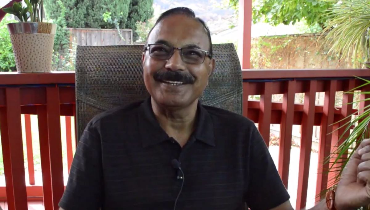 A man wearing glasses is smiling while seated outdoors.