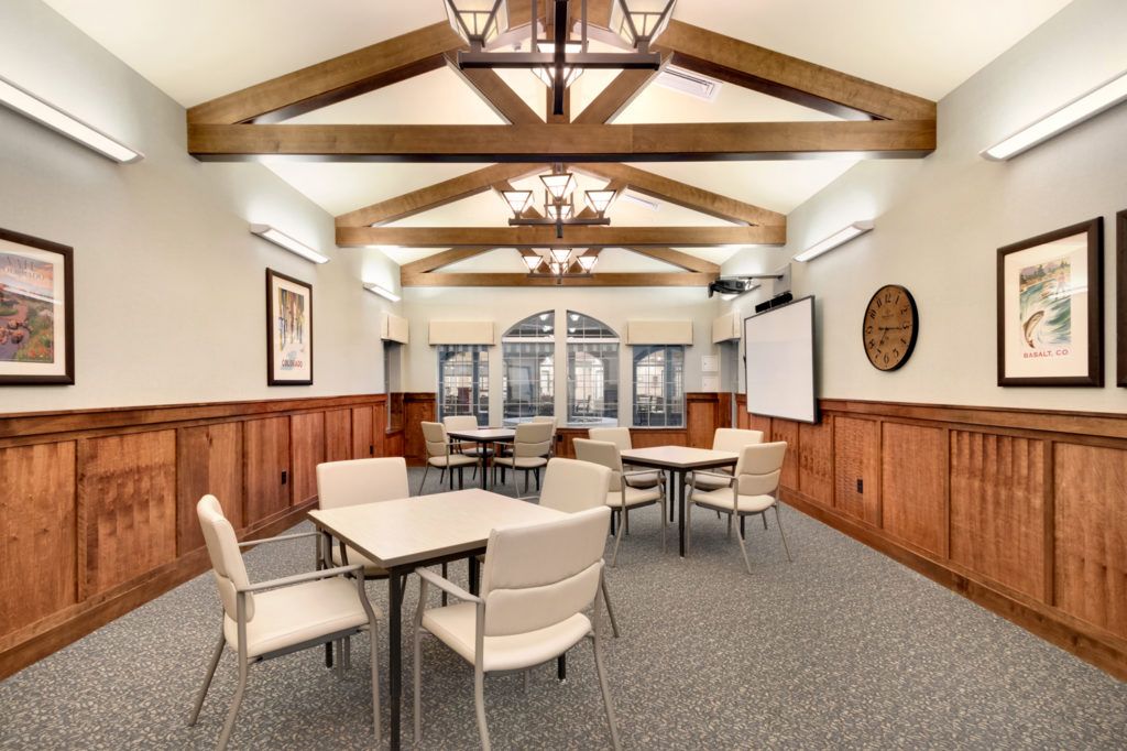 A well-lit meeting room featuring wooden beams, chandeliers, and several tables with chairs arranged in a collaborative setting.