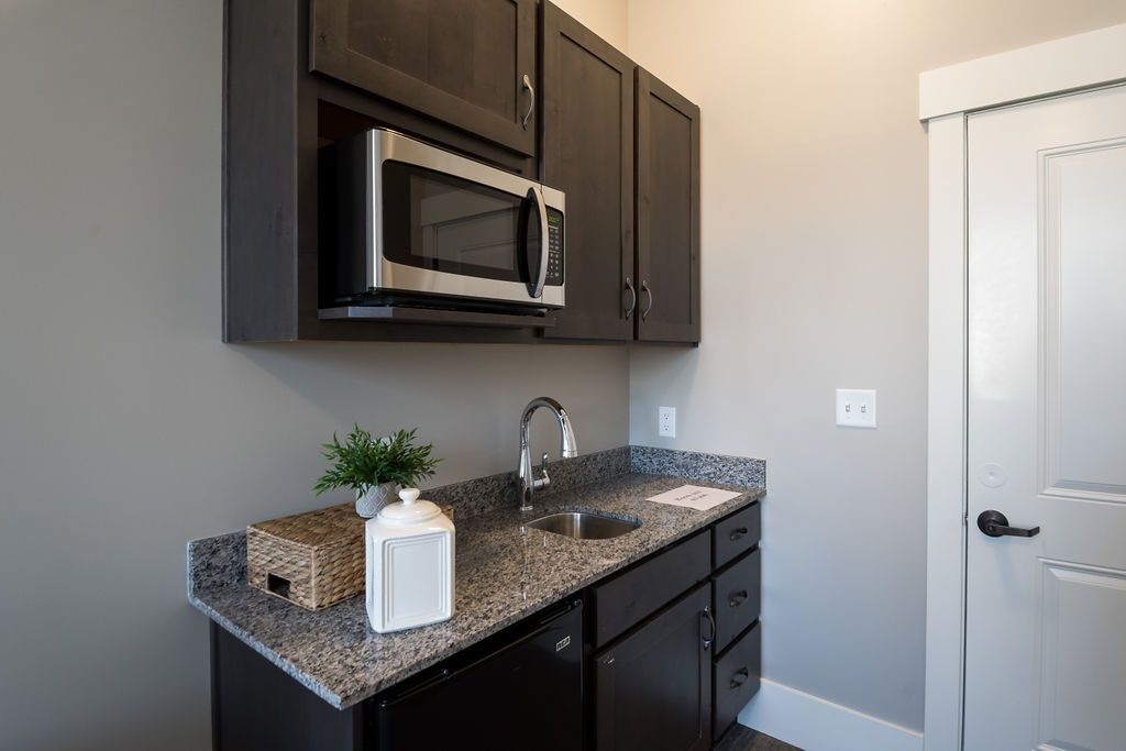 A compact kitchenette featuring dark cabinets, a microwave, and a granite countertop with a sink.