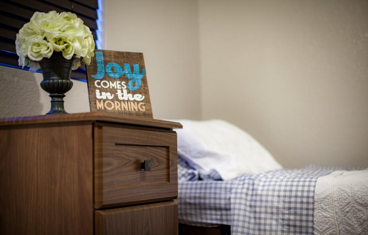 A cozy bedroom with a wooden nightstand, decorated with flowers and a motivational quote sign.