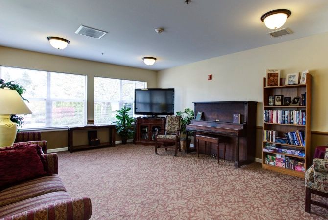 A cozy living room featuring a piano, TV, and bookshelves, with ample natural light from large windows.
