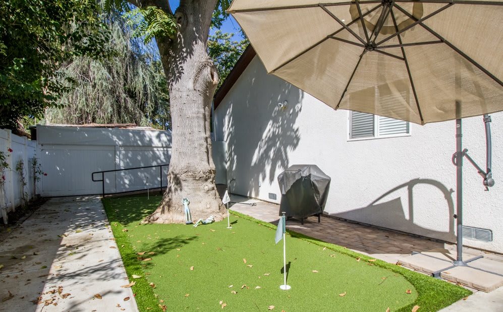 A sunny backyard with a putting green, large tree, patio umbrella, and part of a house.