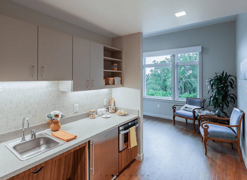 A modern kitchen with wood cabinets and bright lighting opens to a cozy living area with large windows.