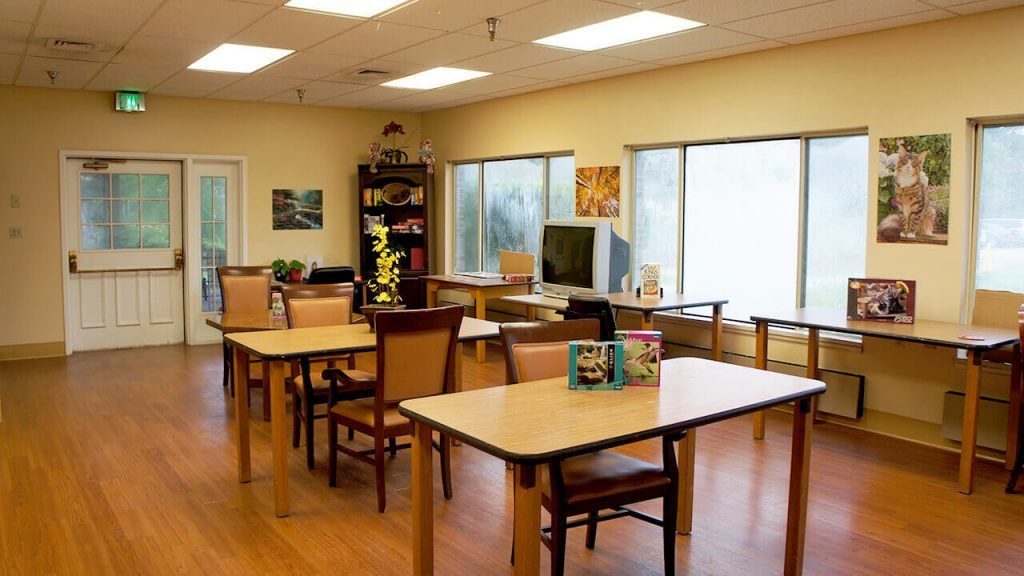 An empty classroom with tables and chairs arranged neatly, with large windows providing natural light.