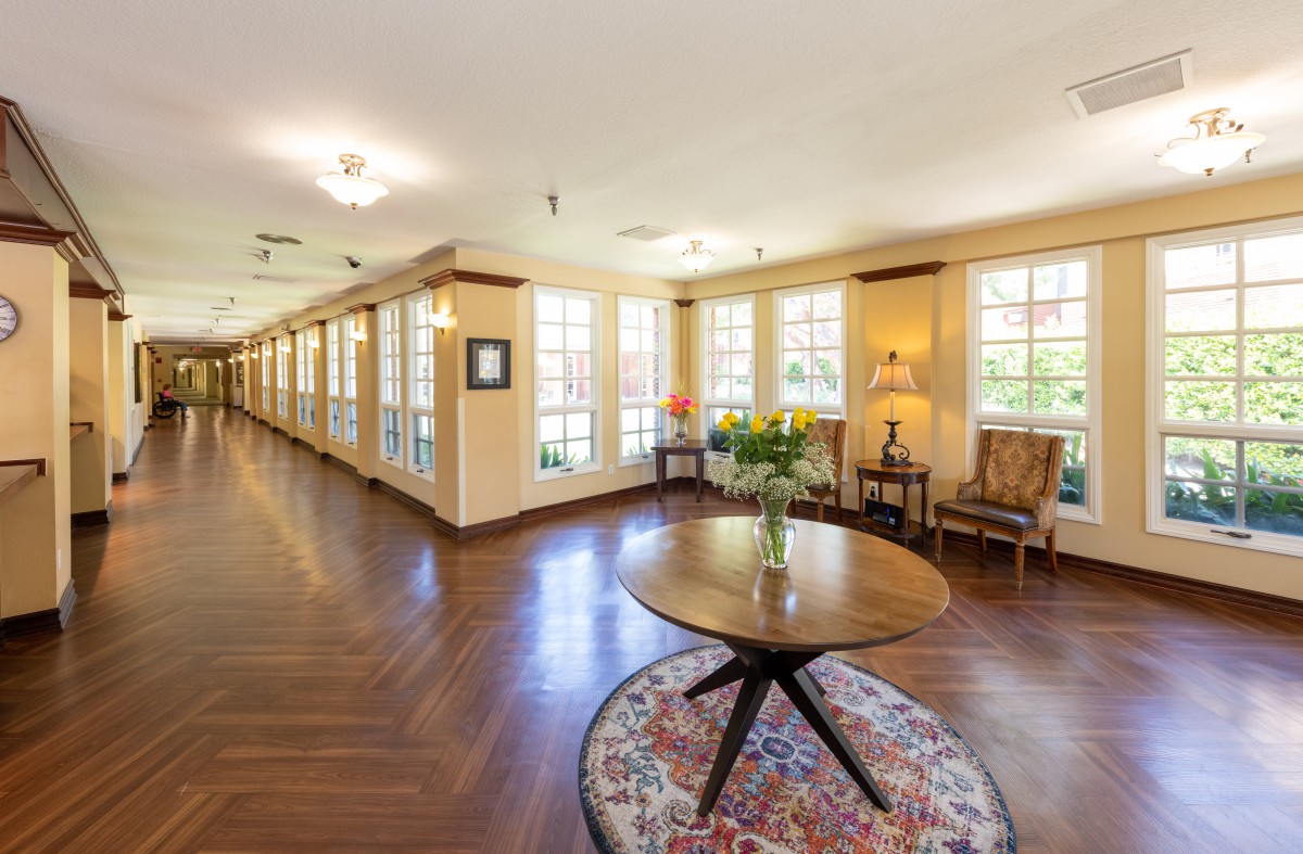 A spacious hallway with wooden floors, featuring a central round table and chairs alongside large windows.