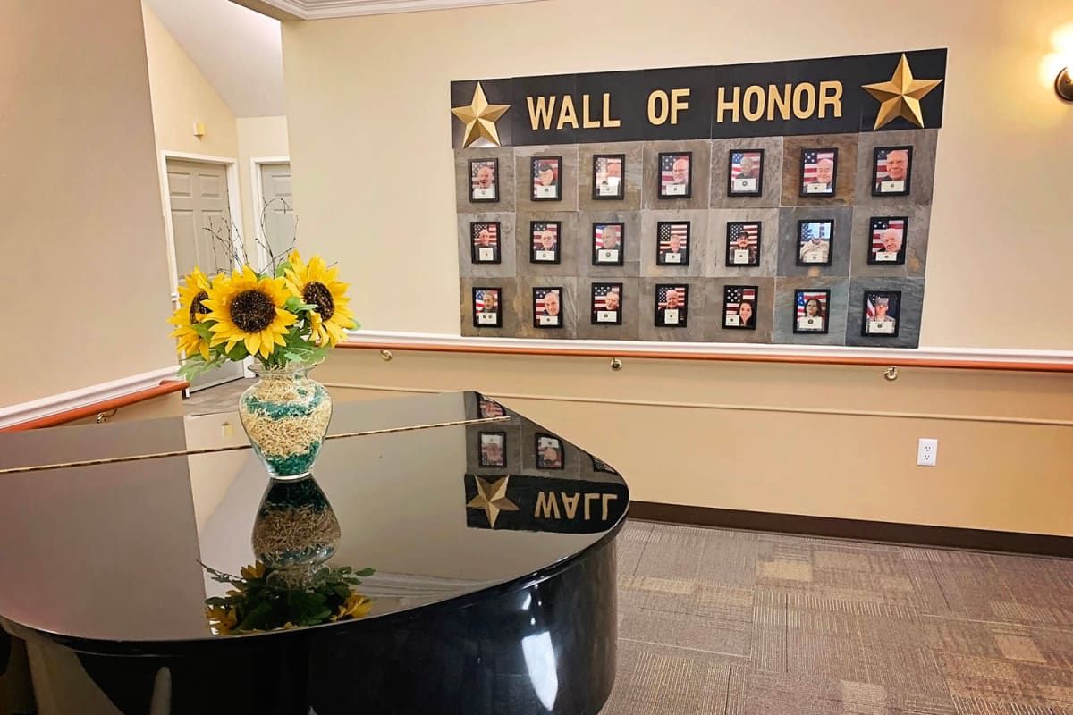 An interior space featuring a 'Wall of Honor' adorned with plaques and a grand piano topped with a vase of sunflowers.