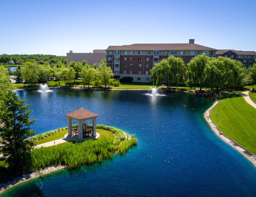 Scenic view of a lakeside area with a gazebo, fountains, and surrounding greenery.