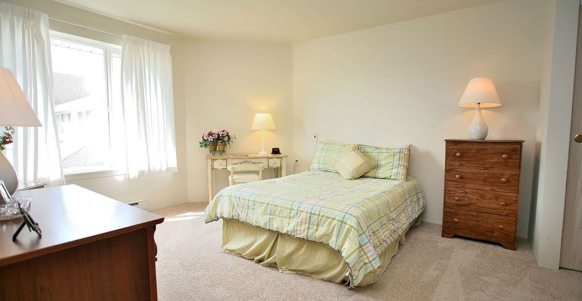 A cozy bedroom with a neatly made bed, nightstand with lamp, and a window letting in natural light.