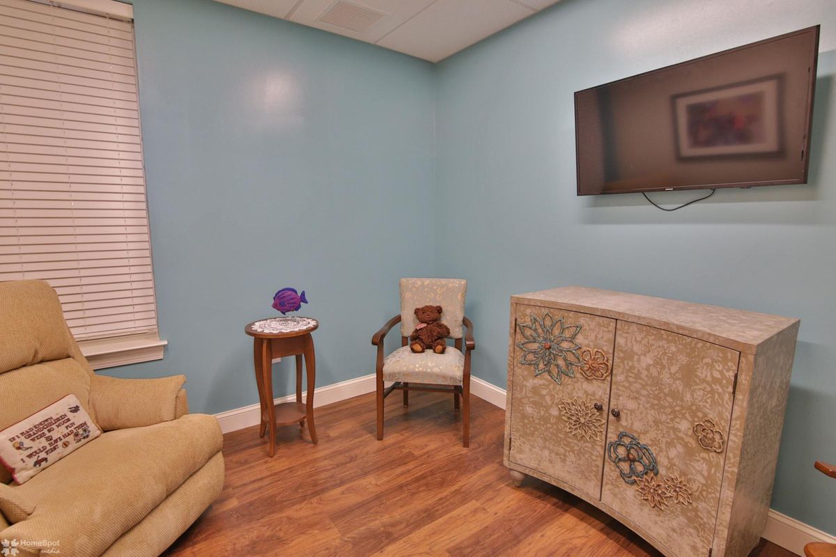 A cozy room with soft blue walls, featuring a chair with a teddy bear, a small round table with a decorative item, an ornate cabinet, and a wall-mounted TV.