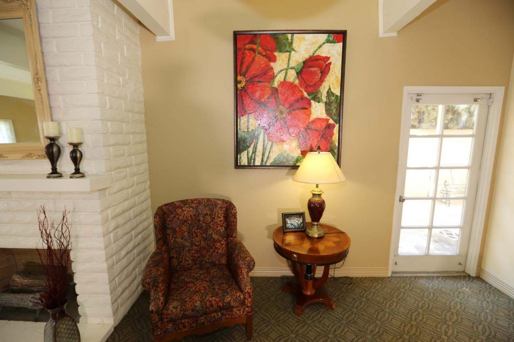 A cozy living room corner with a patterned chair, side table, and a decorative lamp beneath vibrant artwork.