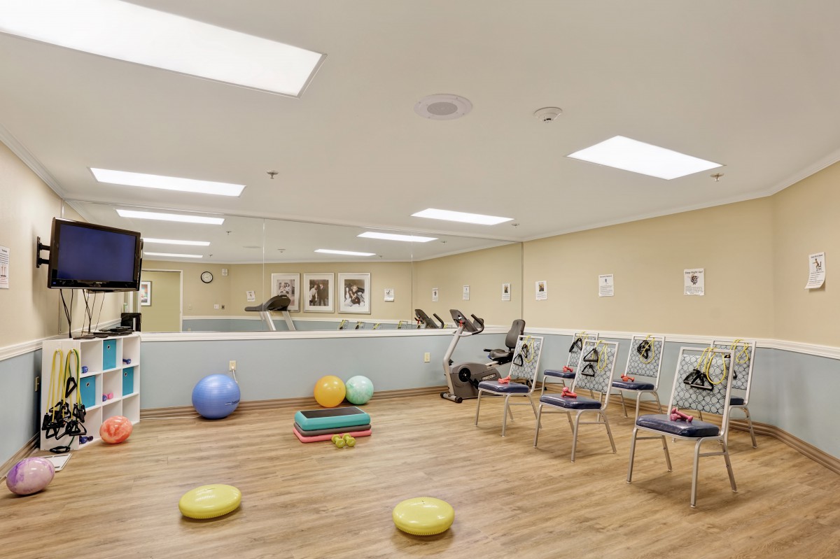 A well-equipped exercise room featuring fitness equipment, stability balls, and chairs arranged on a wooden floor.