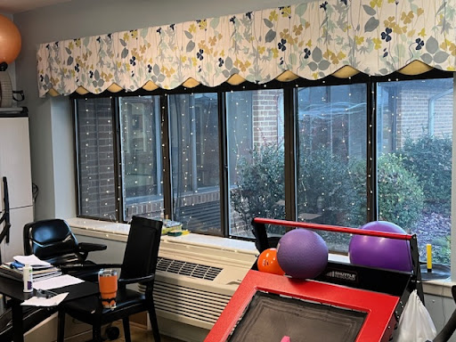 A cozy room with large windows, decorated with floral curtains, featuring a table and chairs along with exercise balls.