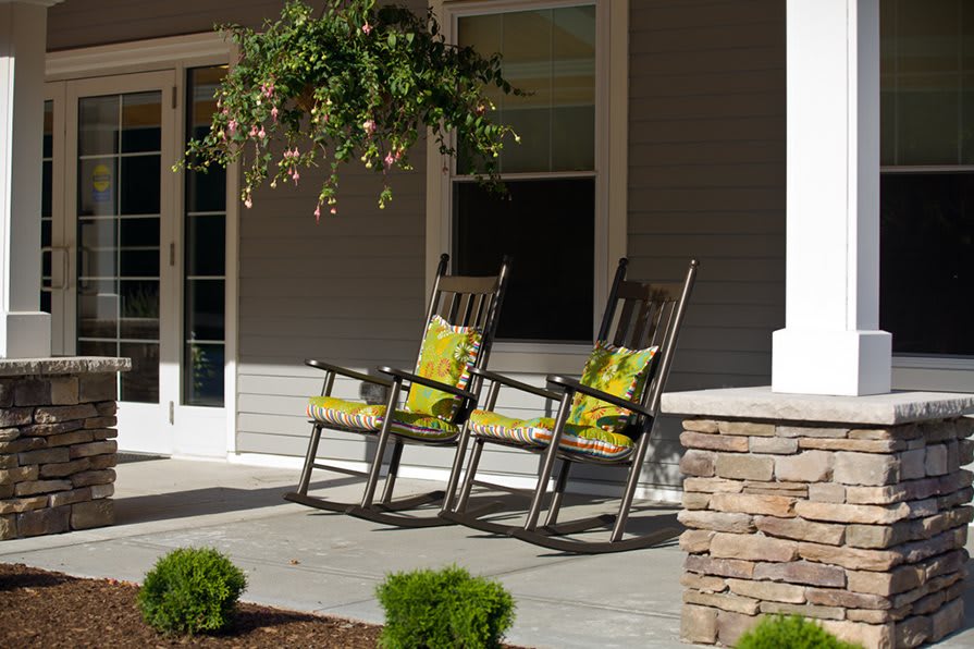 A cozy porch with two rocking chairs adorned with green cushions, surrounded by plants and a hanging basket.