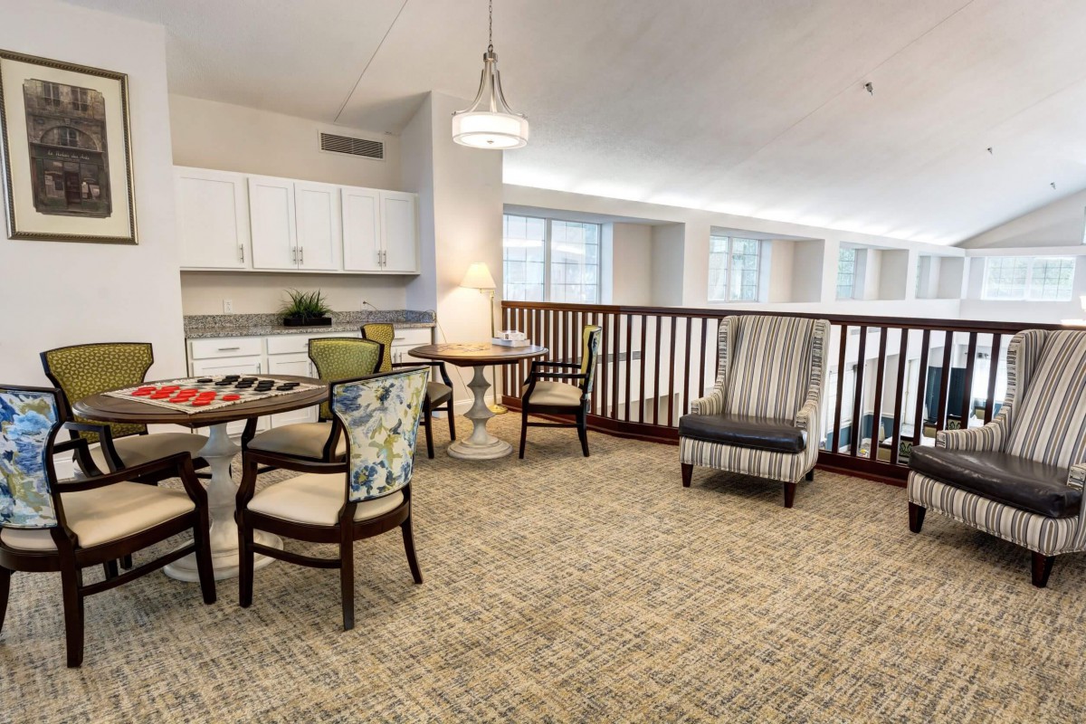 A cozy living area with a table, chairs, and an armchair near a railing overlooking large windows.
