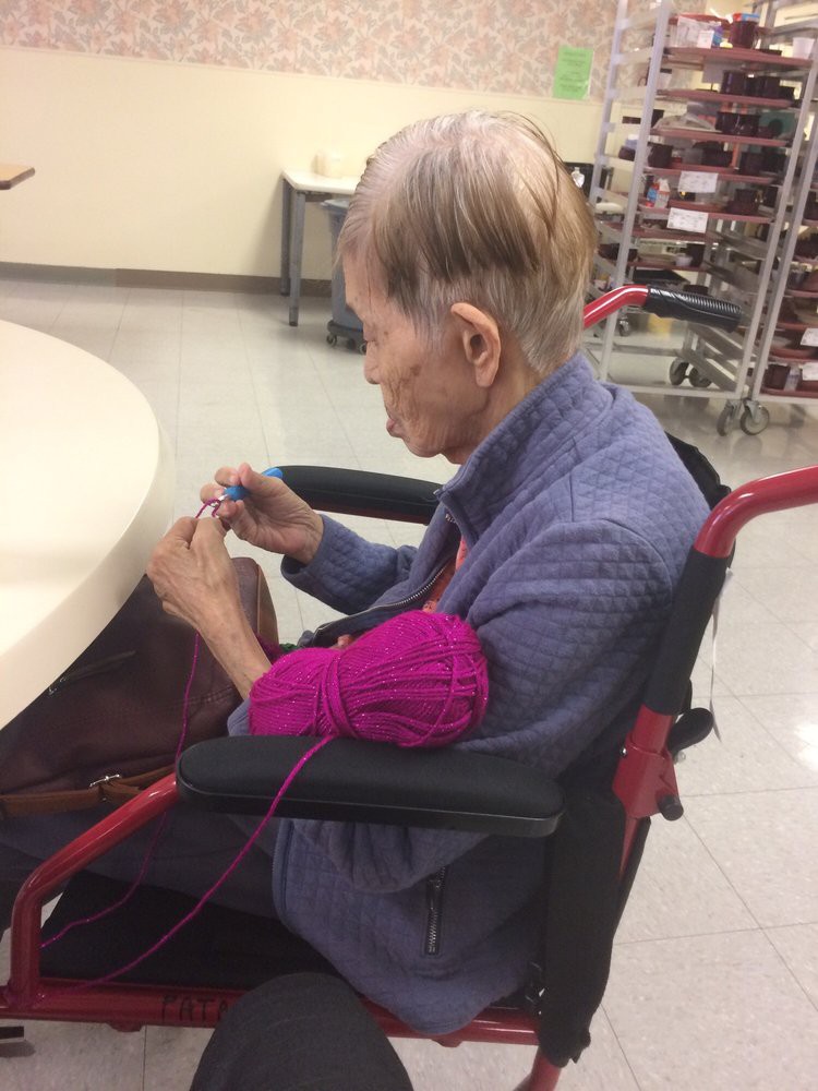 An elderly person in a wheelchair knitting with pink yarn.