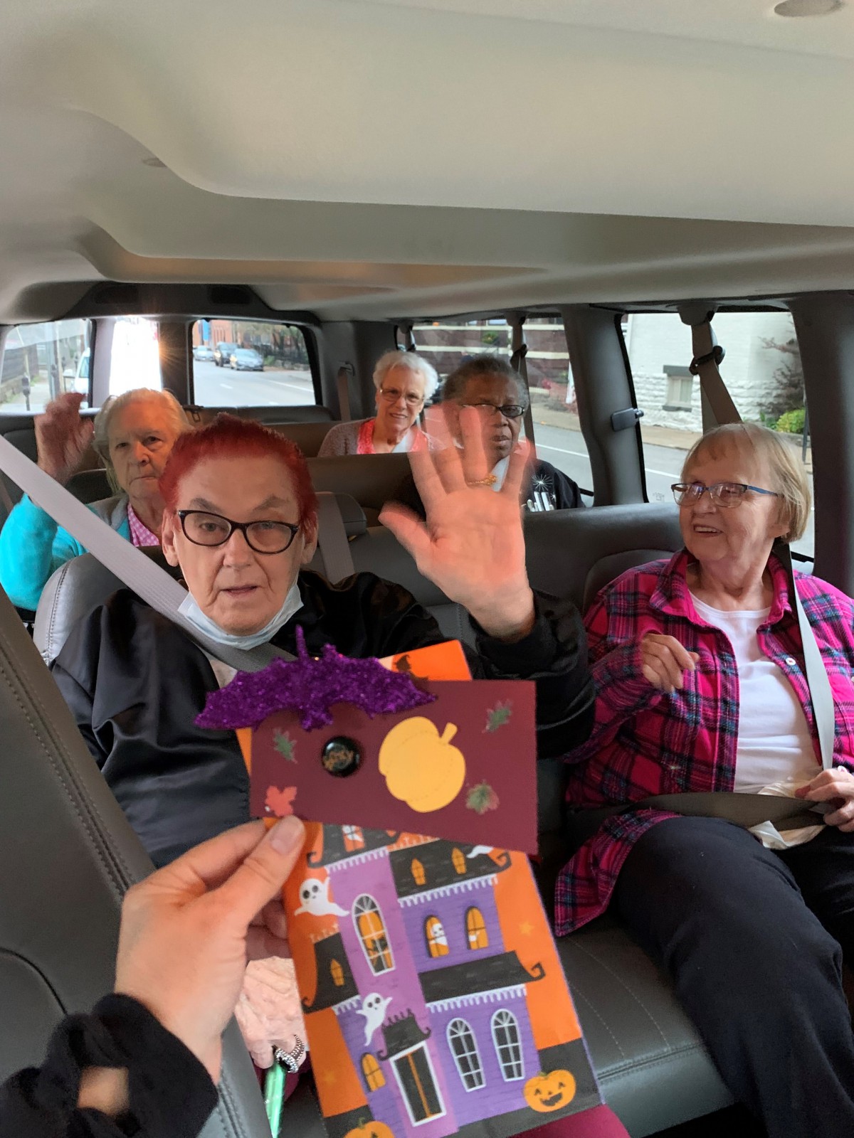 A group of elderly people sitting in a van, with one person waving and greeting the camera.