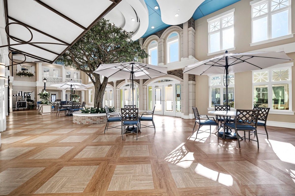 A spacious atrium with seating areas and umbrellas, featuring hardwood flooring and large windows allowing natural light.