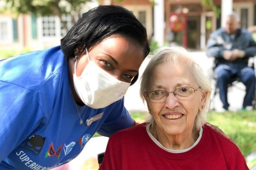 A caregiver wearing a mask posing with an elderly person outdoors, both smiling.