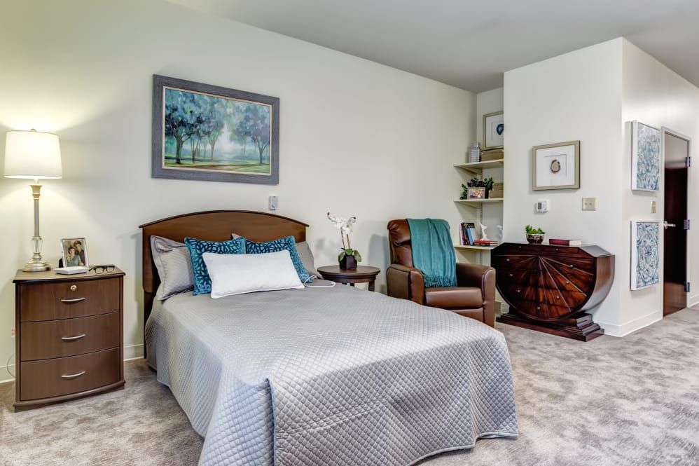 A cozy bedroom featuring a single bed with blue and gray bedding, an armchair with a teal throw, decorative paintings, and carpeted flooring.