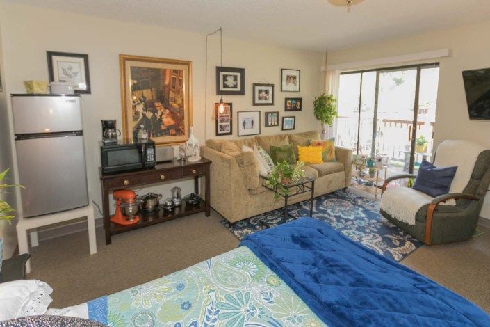 A cozy living room with a beige sofa, framed artwork and photos on the wall, a coffee table, and a sliding glass door leading outside.