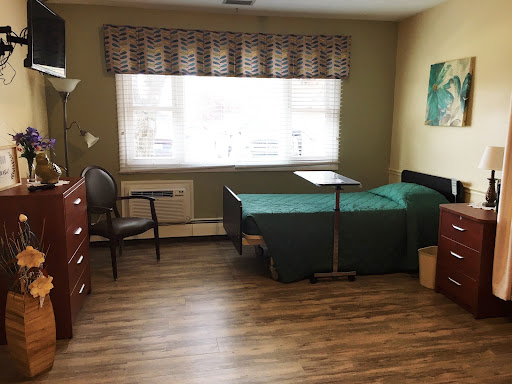 A well-lit, simple room featuring a bed, chair, and dresser, with a view through the window.