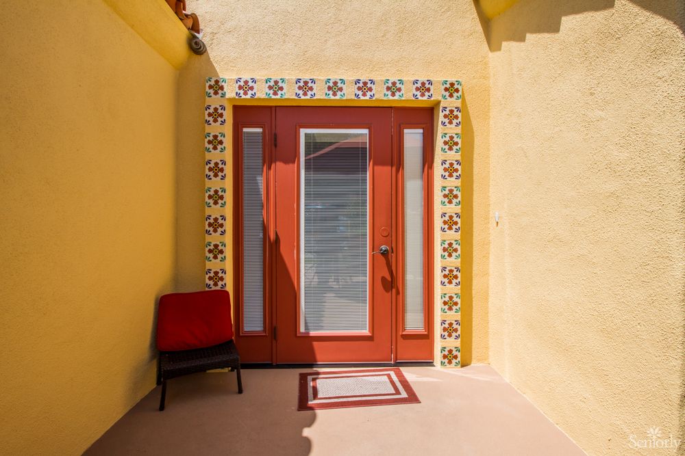 A sunlit entrance with a red door framed by patterned tiles, set against a vibrant yellow wall and accompanied by a chair.