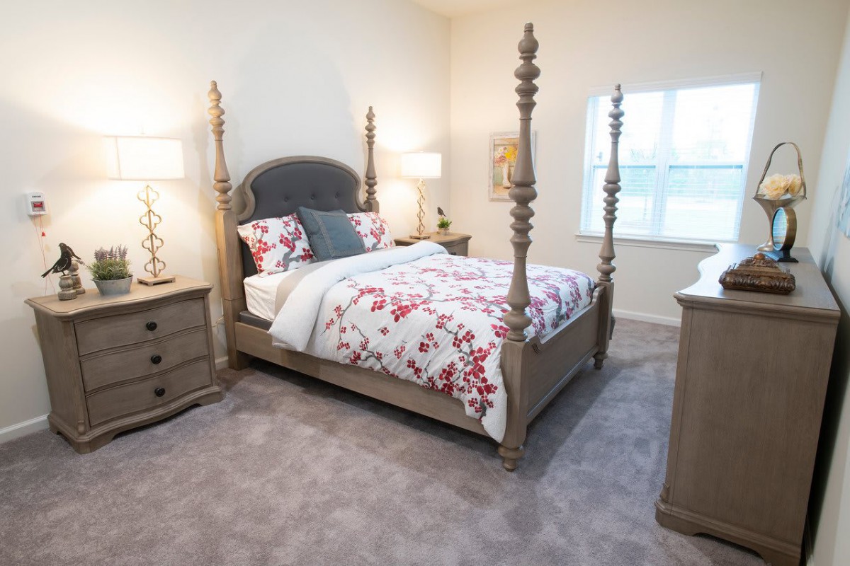 A cozy bedroom featuring a four-poster bed with floral bedding, complemented by two nightstands and table lamps, on a carpeted floor.