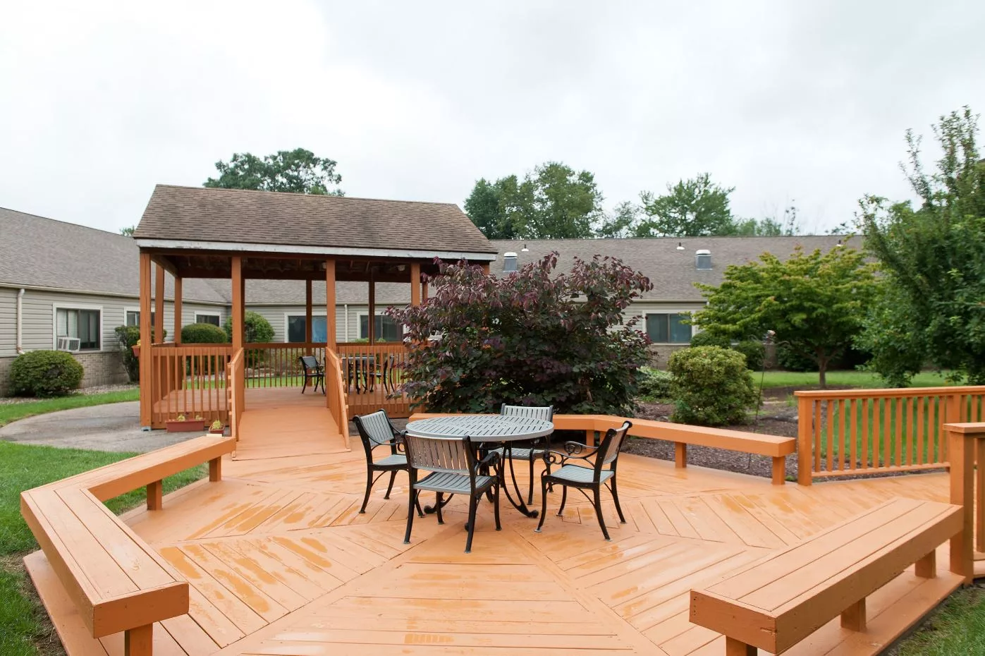 An outdoor deck area featuring a central table with chairs, surrounded by lush greenery and a gazebo.