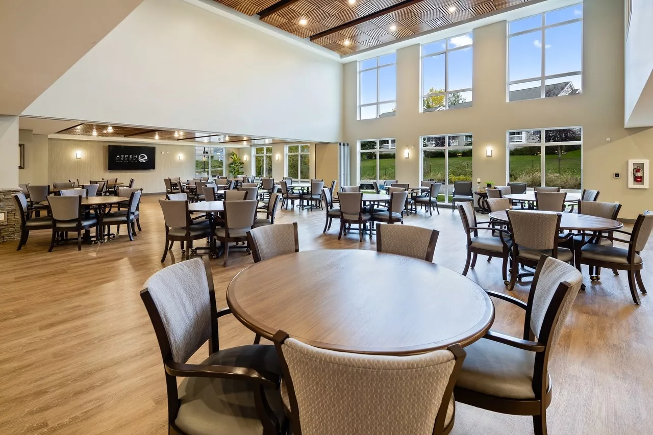 A spacious dining area with multiple tables and chairs, featuring large windows and wooden flooring.