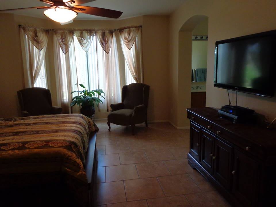 A cozy bedroom with a bed, armchairs, TV, and natural lighting from large windows.