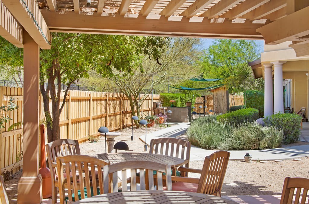 A cozy backyard patio with wooden chairs and table set under a pergola, surrounded by lush greenery and a well-maintained garden.