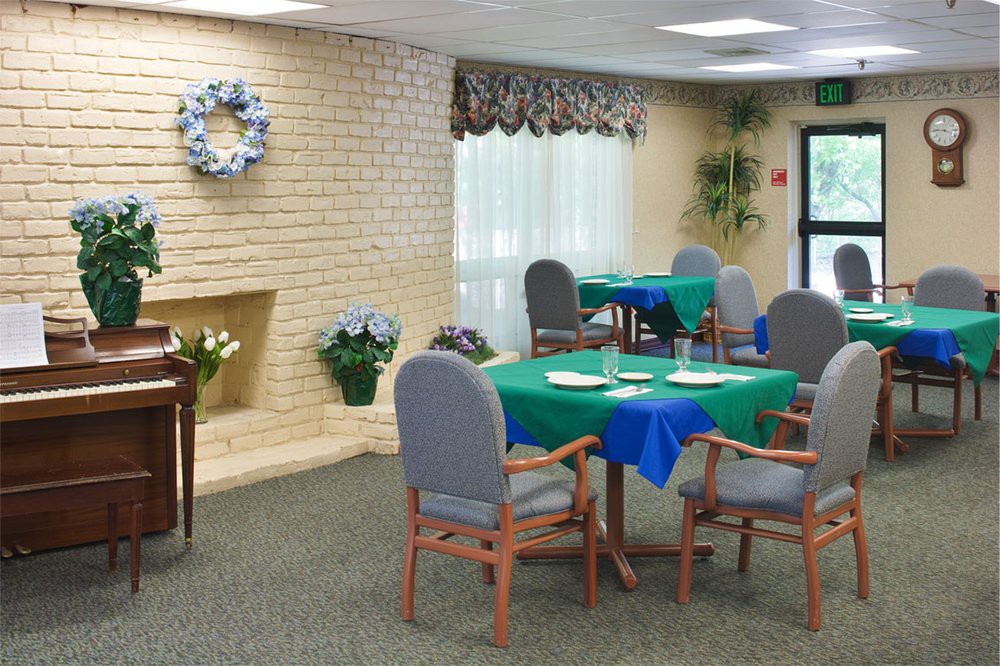 A cozy dining area with tables and chairs, featuring green and blue tablecloths, indoor plants, and natural lighting through large windows.