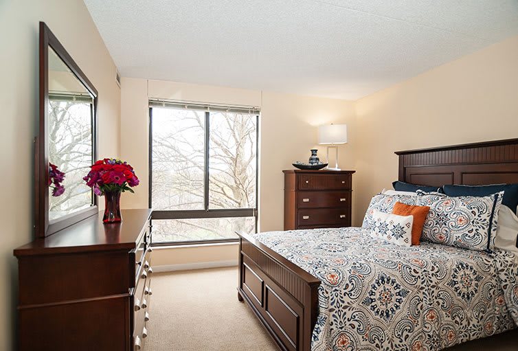 A cozy bedroom with wooden furniture and a large window, featuring a neatly made bed and decorative elements.