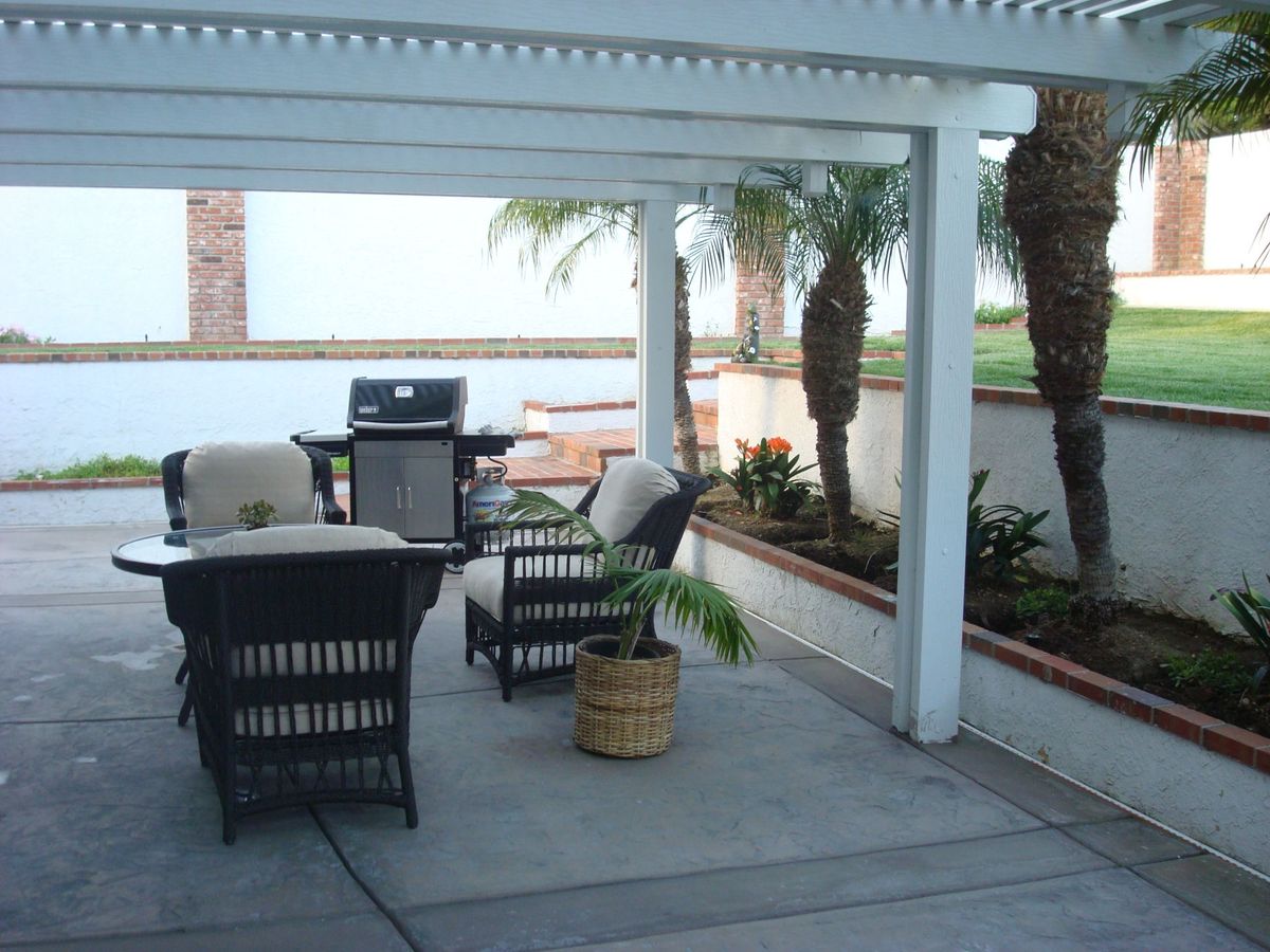 A cozy outdoor patio with a covered pergola, featuring seating and a grill, surrounded by palm trees.