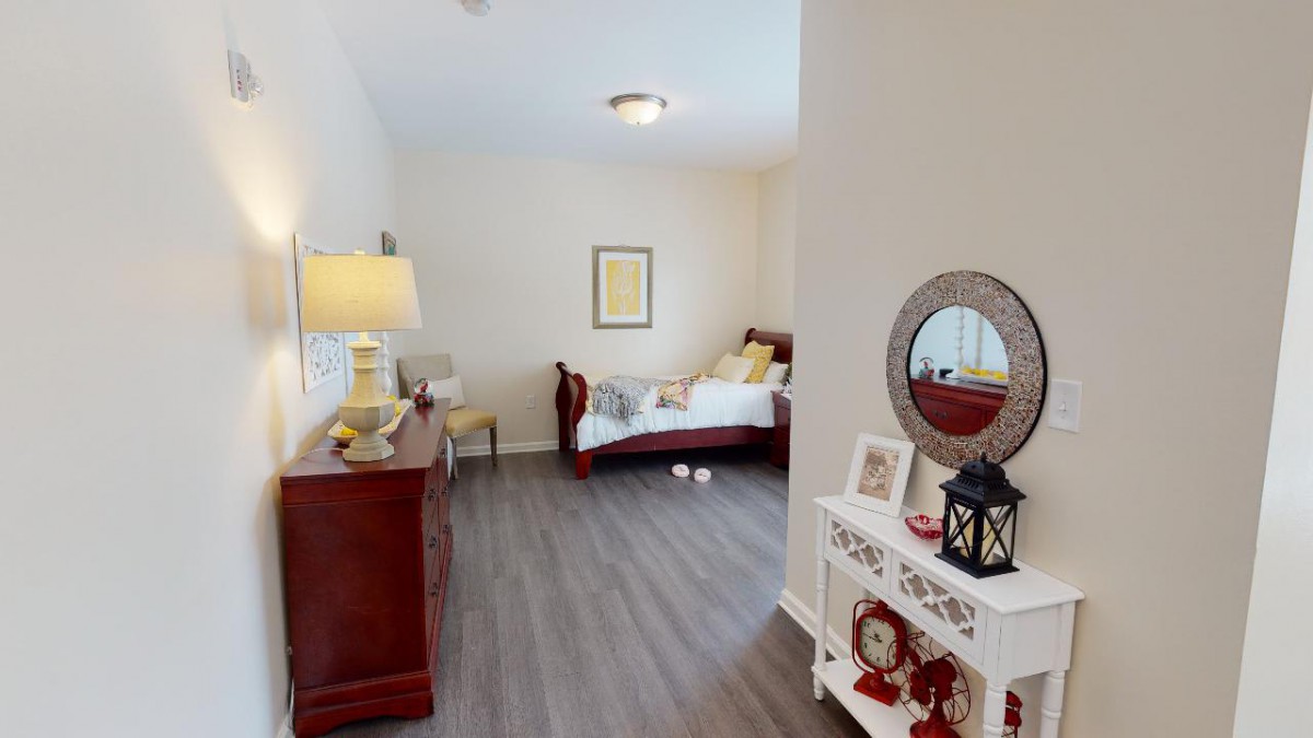 A cozy bedroom with wooden flooring, featuring a bed, dresser, and decorative elements like a lamp and a mirror.