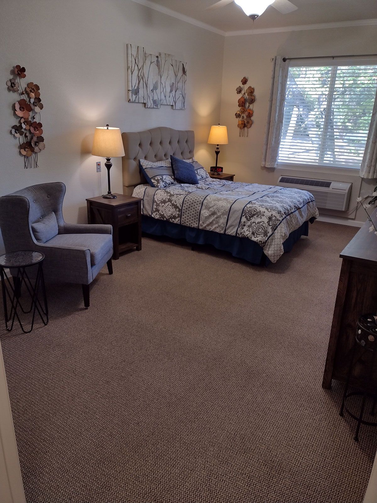 A cozy bedroom with a bed, chair, and bedside lamps, featuring wall decor and a large window.
