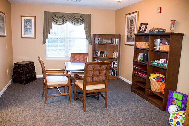 A cozy office space with a table and chairs, surrounded by bookshelves and natural light from the window.