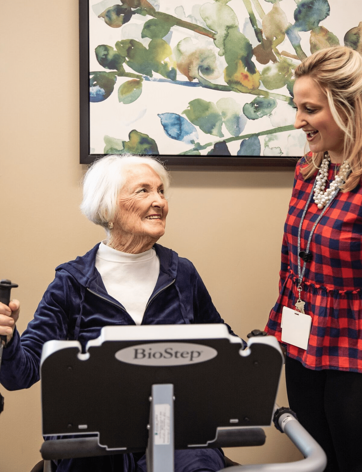 An elderly woman exercises on a machine while interacting with a trainer indoors.