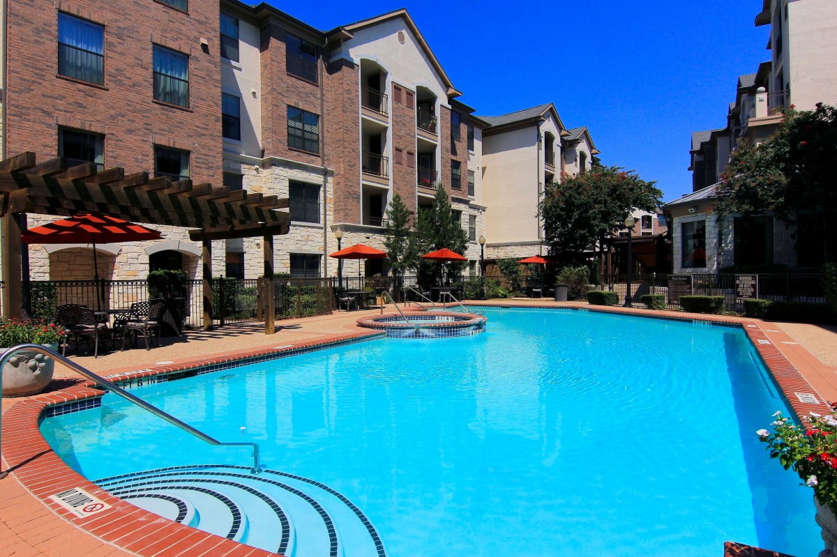 A bright outdoor swimming pool area in a residential apartment complex with clear blue skies overhead.