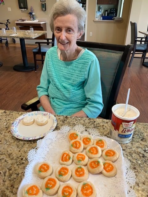 An elderly woman smiles while sitting at a table with a plate of cookies and a drink.