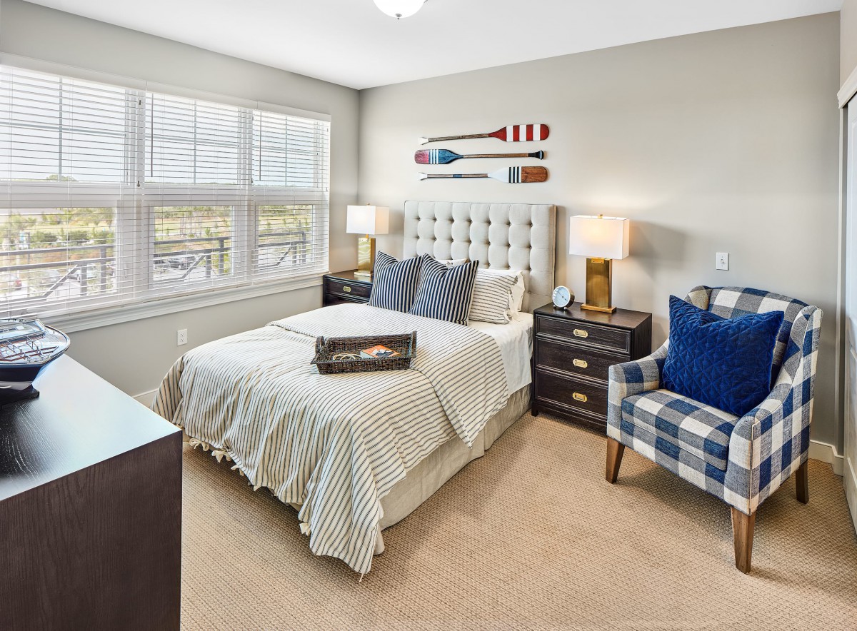 A modern, cozy bedroom featuring a neatly made bed with patterned bedding, a tufted headboard, and bedside tables with lamps. A comfortable chair with blue accents is positioned next to the window, allowing natural light to brighten the room.