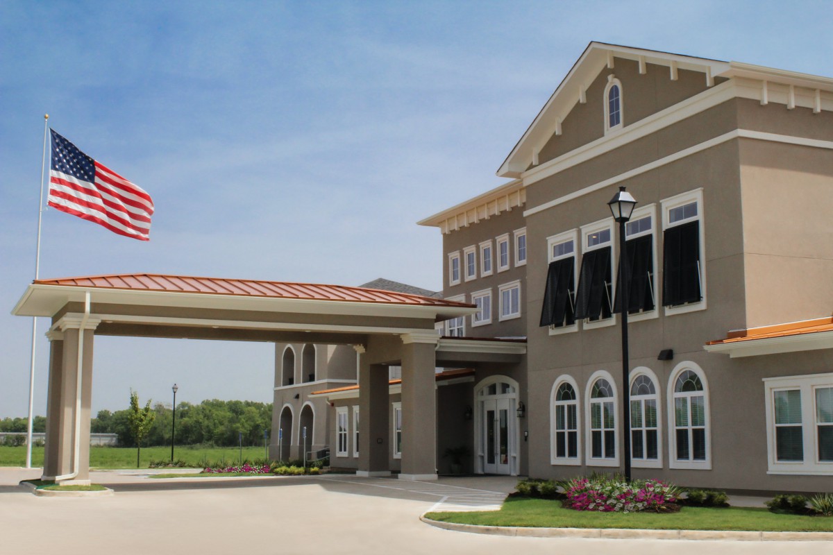 A modern building with an entrance canopy and an American flag, surrounded by landscaped gardens on a sunny day.