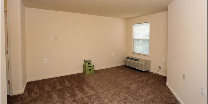 An empty room with brown carpet, a window, two green boxes, and an air conditioner.