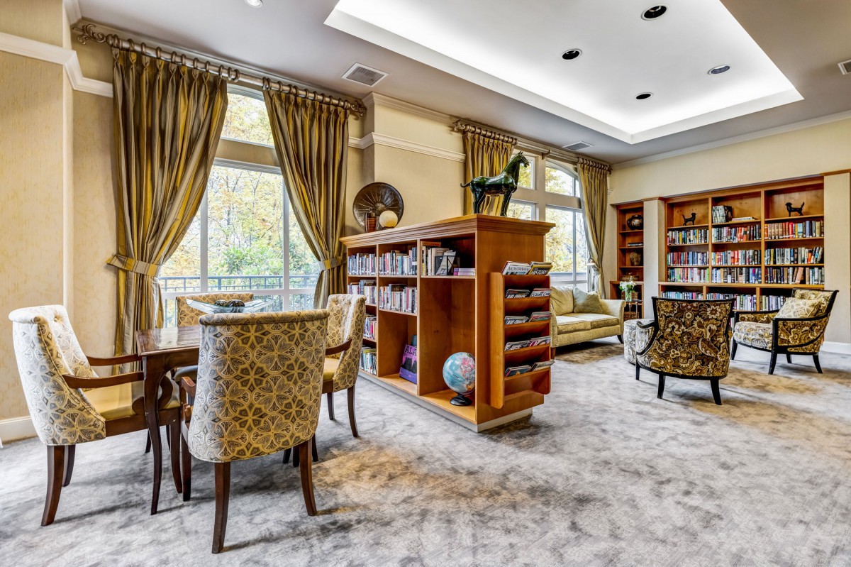 A spacious and elegant living room with a library section, featuring bookshelves, comfortable seating, large windows with curtains, and a carpeted floor.