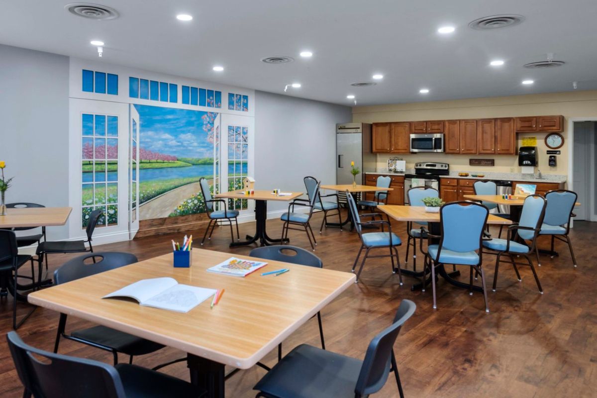 A well-lit classroom with multiple tables and chairs, featuring a kitchen area and a vibrant mural on the wall.