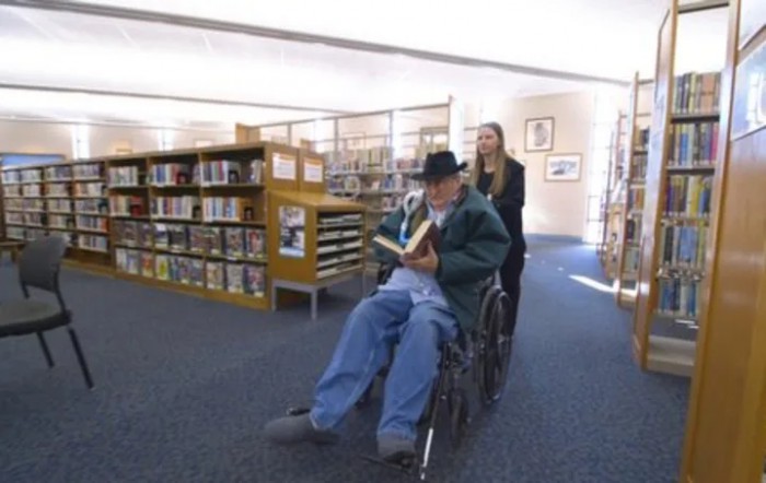 A person in a wheelchair is being pushed by another individual in a library, reading a book.